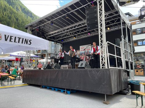 A band is playing on a stage in a festival atmosphere. Guests are lingering, enjoying the music under a clear sky.