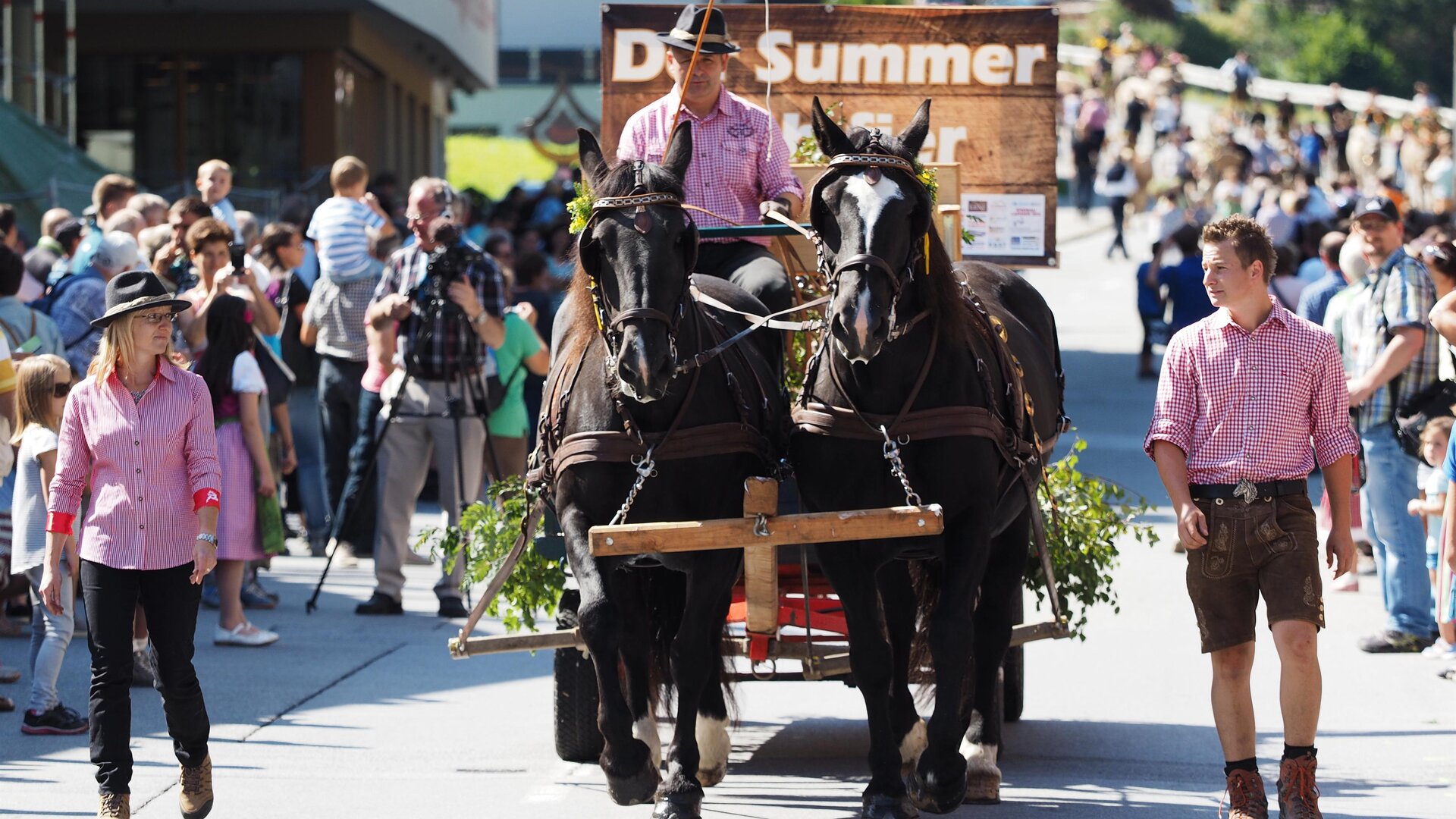 A horse-drawn wagon moves through a busy street. Many spectators in traditional clothing stand along the way and watch the parade.