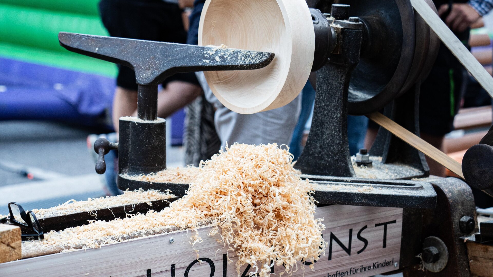 A woodworking machine processes wood into a bowl. Wood shavings are lying on the table and there are people visible in the background.