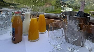 A table with various drinks, including juices and sparkling wine, stands in front of an impressive mountain landscape. The clouds gently drift over the green hills.