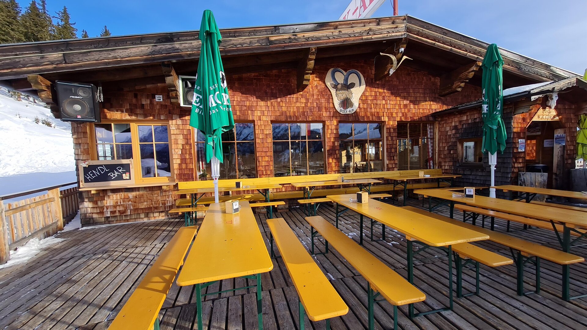 A rustic mountain cabin with a wooden terrace and yellow benches. There are green umbrellas and a snowy landscape in the background.
