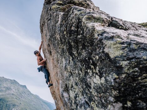 A climber ascends a large rock while her partner supports her. In the background, impressive mountains and a blue sky can be seen.