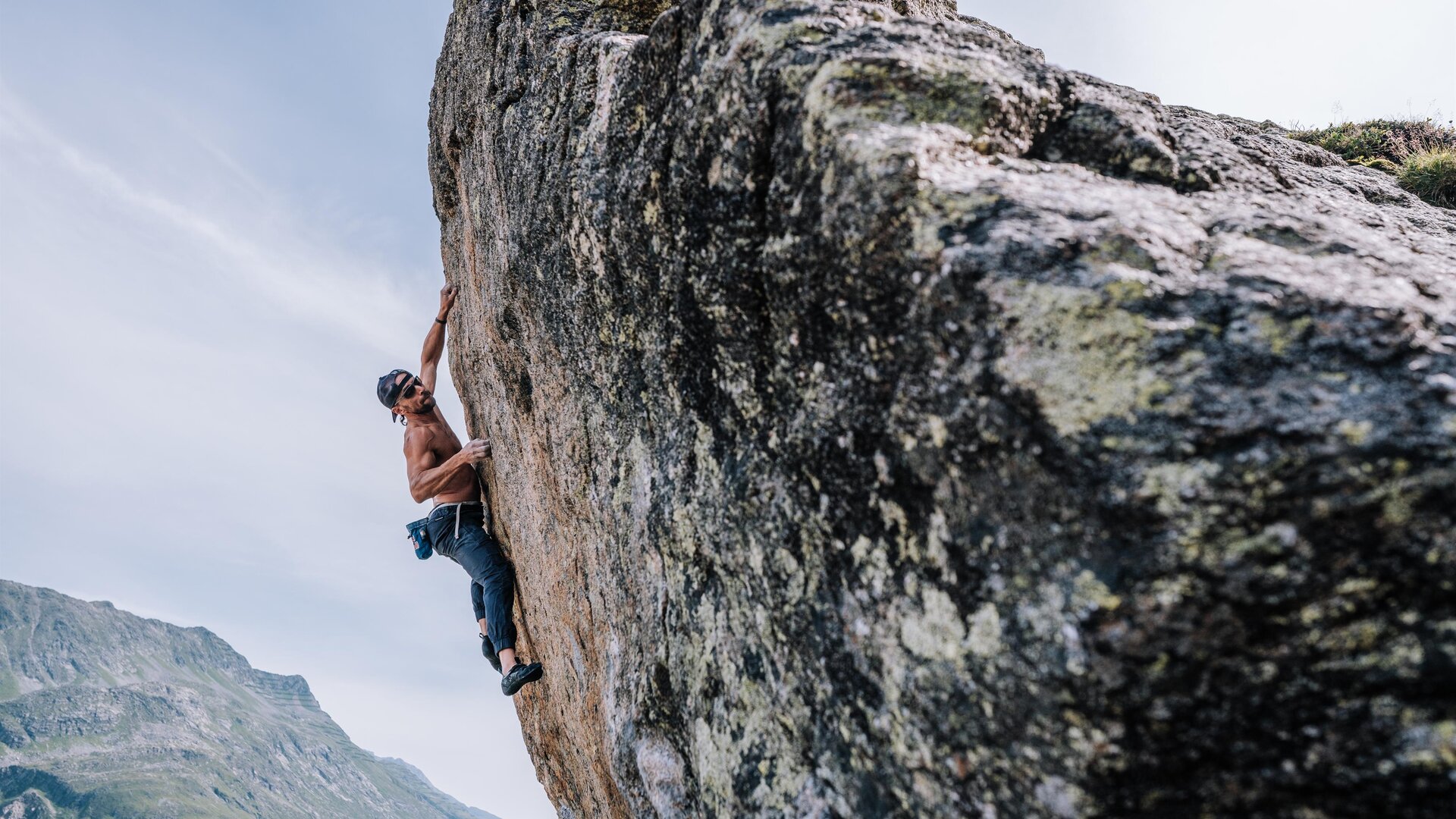 A climber ascends a large rock while her partner supports her. In the background, impressive mountains and a blue sky can be seen.