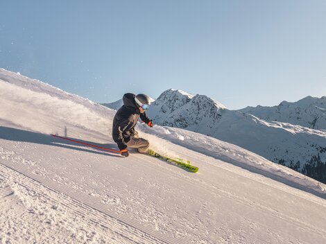 Ein Skifahrer fährt die Piste hinunter. Im Hintergrund sind schneebedeckte Berge und ein klarer Himmel zu sehen.