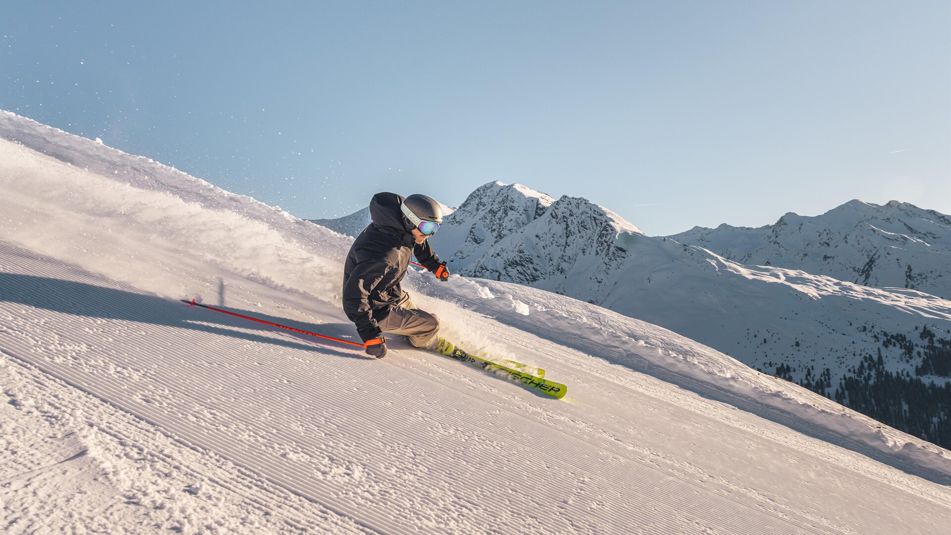 Ein Skifahrer fährt die Piste hinunter. Im Hintergrund sind schneebedeckte Berge und ein klarer Himmel zu sehen.