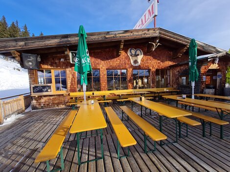 A cozy mountain cabin with wooden cladding and several long benches. The terrace is equipped with green sun umbrellas and the snow is visible in the background.