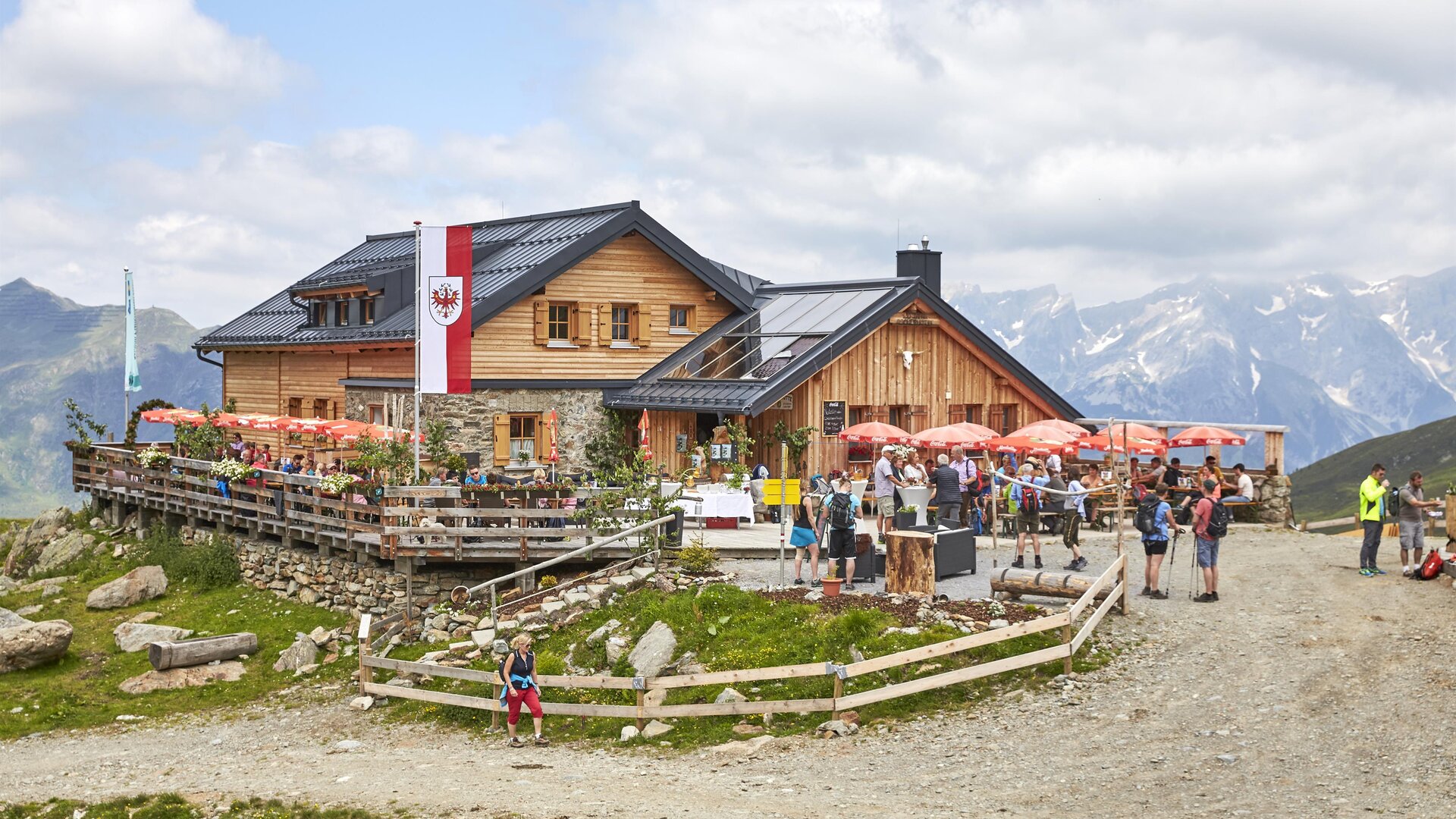 Eine gemütliche Berghütte mit einer Terrasse und vielen Besuchern. Im Hintergrund sind hohe Berge und ein teilweise bewölkter Himmel zu sehen.