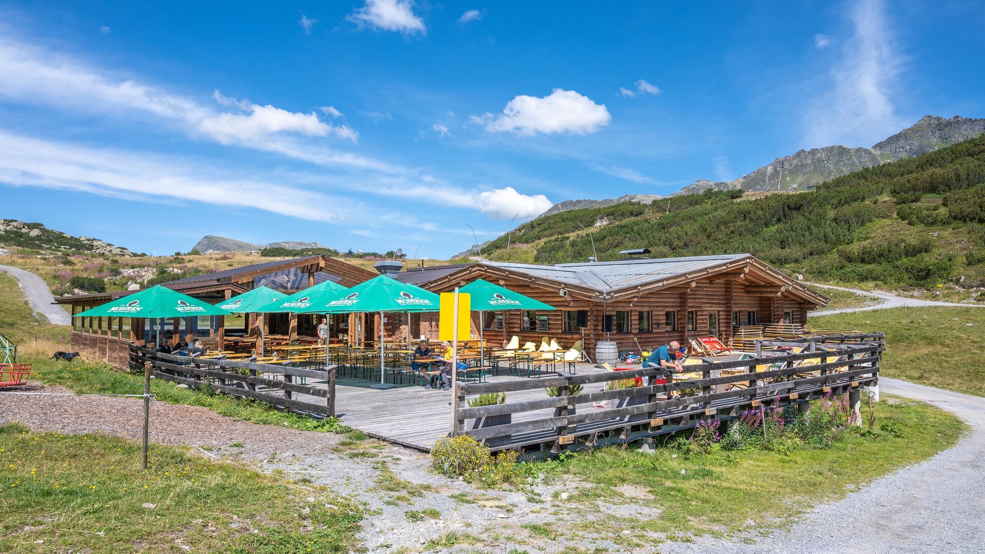A cozy wooden house with a terrace and green sunshades. The surroundings are mountainous and green under a blue sky.