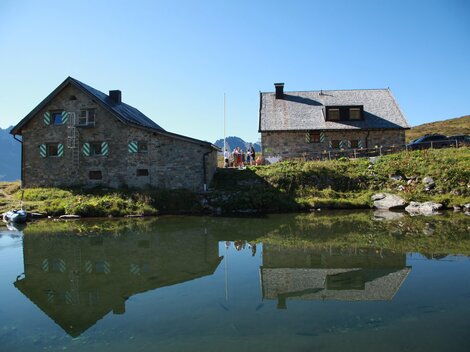 A picturesque mountain landscape with two cozy houses along a tranquil lake. The buildings reflect clearly in the water and are surrounded by green meadows.