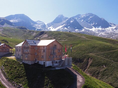 A modern wooden building is situated picturesque in the mountains. In the background, snow-covered peaks and a green landscape can be seen.