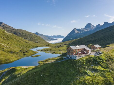A modern wooden building is situated picturesque in the mountains. In the background, snow-covered peaks and a green landscape can be seen.