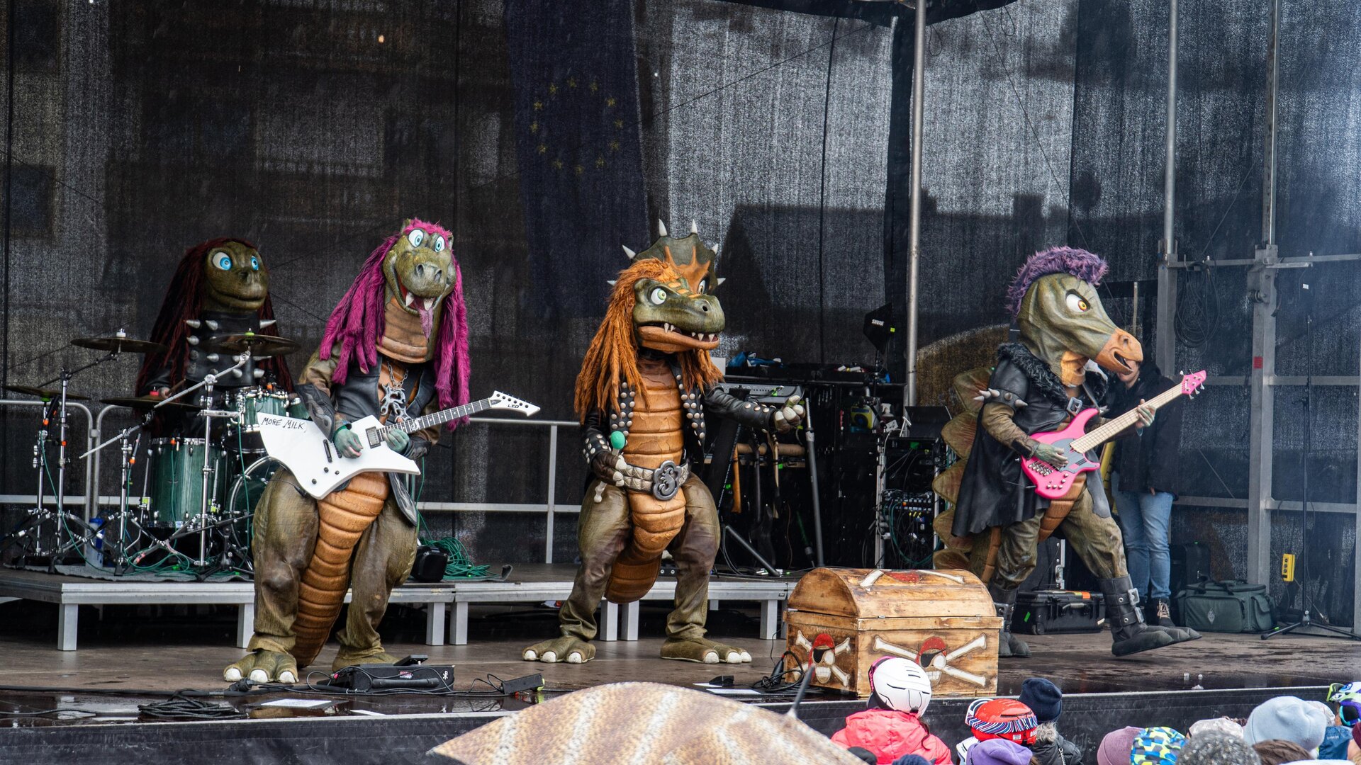 A band of dragon mascots is playing on a stage. In the background, musical instruments and an enthusiastic crowd can be seen.