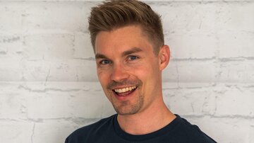 A young man with short, blond hair smiles kindly. He is wearing a black T-shirt with the print "ALPHATAURI".