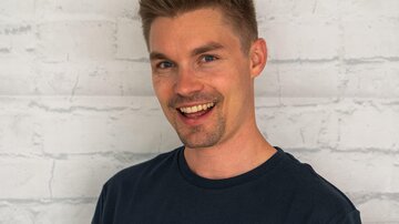 Ein junger Mann mit kurzem, blondem Haar lächelt freundlich. Er trägt ein schwarzes T-Shirt mit dem Aufdruck "ALPHATAURI".