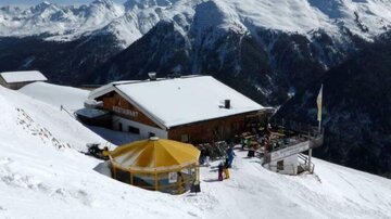 Ein Restaurant in den Bergen, umgeben von schneebedeckten Gipfeln. Die sonnige Terrasse bietet einen schönen Ausblick auf die winterliche Landschaft.