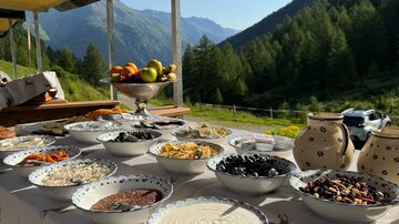 Ein Tisch mit verschiedenen Schalen voller Zutaten für ein Buffet steht in einer Berglandschaft. Im Hintergrund sind grüne Berge und ein klarer Himmel zu sehen.