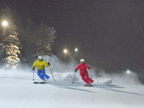 Zwei Skifahrer fahren nachts auf einer schneebedeckten Piste. Die Umgebung ist beleuchtet und erzeugt eine winterliche Atmosphäre.