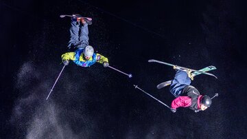 Two skiers are performing acrobatic jumps in the air. The background is dark, and it is visible how snow is being kicked up.