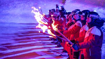A group of skiers in red suits stands on a lit slope with torches. The flames illuminate the winter landscape and create a festive atmosphere.