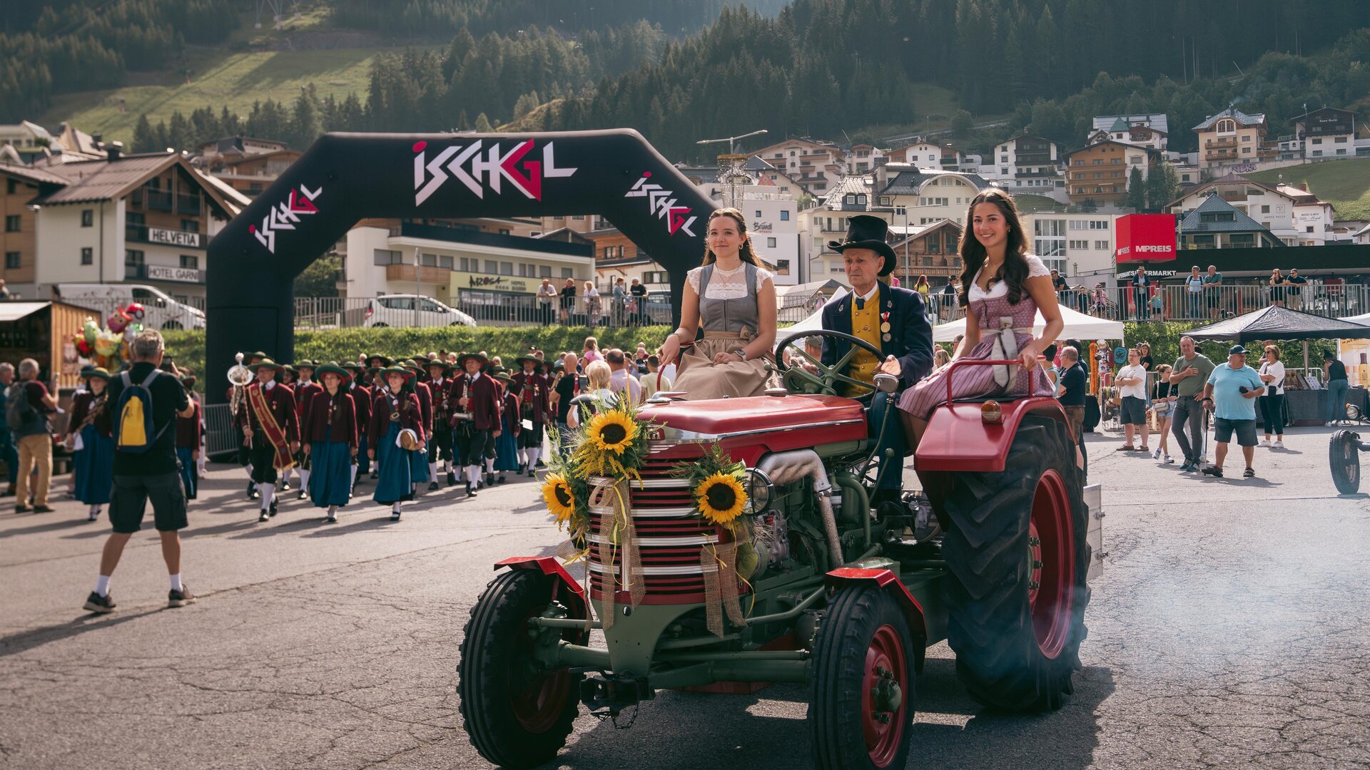 Eine lebhafte Veranstaltung mit einem Traktor, der von zwei Frauen in traditionellen Trachten begleitet wird. Im Hintergrund sind jubelnde Menschen und eine malerische Berglandschaft zu sehen.