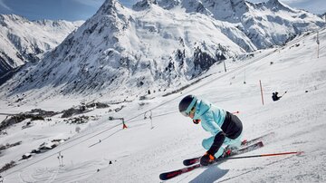 Ein Skifahrer fährt den Hang hinunter, umgeben von schneebedeckten Bergen. Die sonnige Szene zeigt klare blaue Himmel und eine lebendige Winterlandschaft.