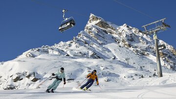 Zwei Skifahrer fahren auf einer schneebedeckten Piste unter einem klaren blauen Himmel. Im Hintergrund sieht man einen Berg und eine Gondelbahn.