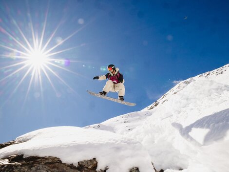 Ein Snowboarder springt über einen schneebedeckten Hügel unter einem strahlend blauen Himmel. Die Sonne scheint hell und schafft eine dynamische Atmosphäre.