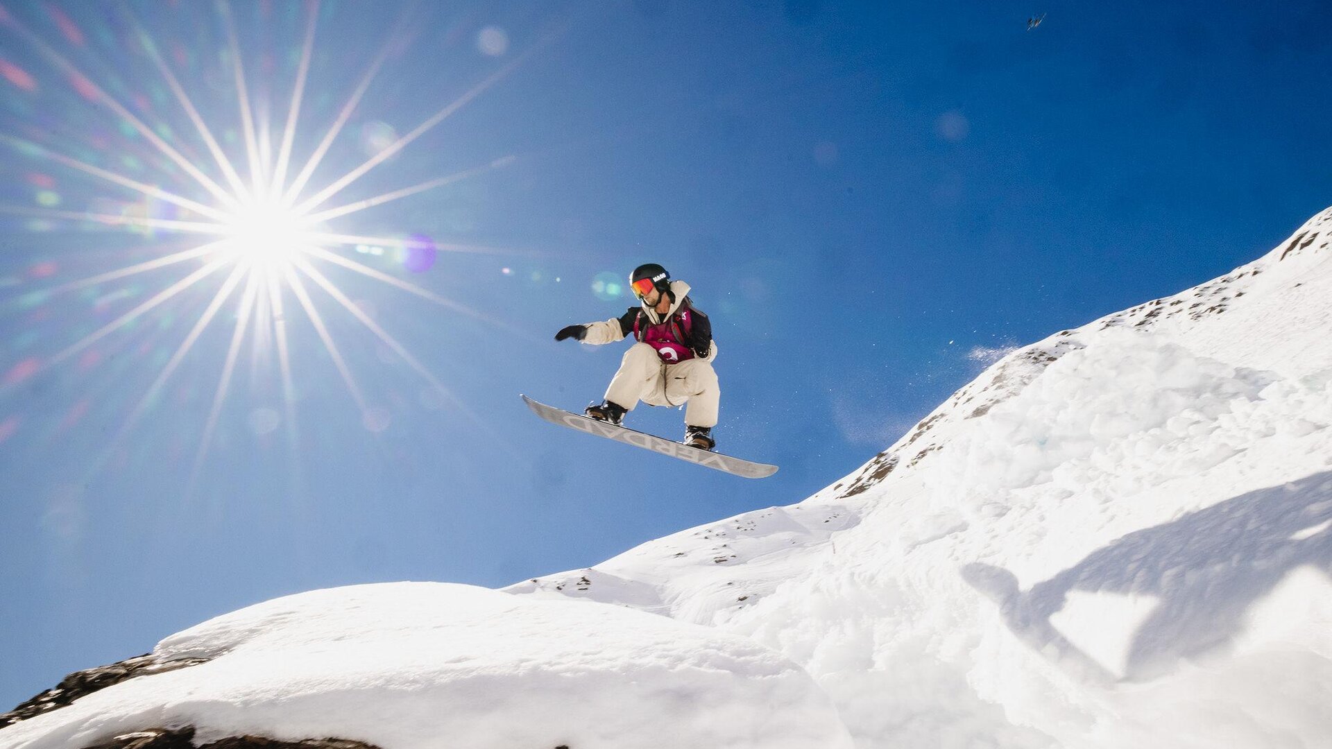 Ein Snowboarder springt über einen schneebedeckten Hügel unter einem strahlend blauen Himmel. Die Sonne scheint hell und schafft eine dynamische Atmosphäre.