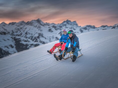 Zwei Personen sitzen auf Schlitten und genießen den Schnee auf einem Berg. Im Hintergrund sind schneebedeckte Berge und ein schöner Sonnenuntergang zu sehen.