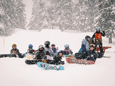 Eine Gruppe von Snowboardern sitzt im Schnee während eines Schneesturms. Um sie herum sind verschneite Bäume und eine winterliche Landschaft.