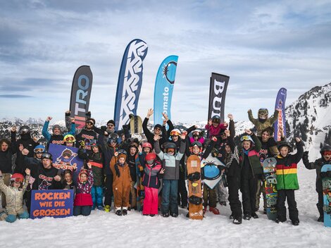 Eine Gruppe von Kindern und Erwachsenen steht fröhlich im Schnee. Sie tragen Skiausrüstung und feiern mit Fahnen im Hintergrund.