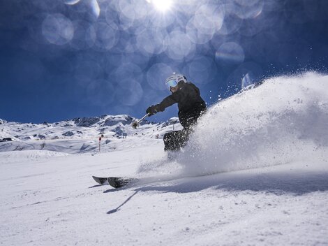 Ein Skifahrer fährt mit Schwung durch den frischen Schnee. Im Hintergrund sind schneebedeckte Berge und ein klarer blauer Himmel zu sehen.