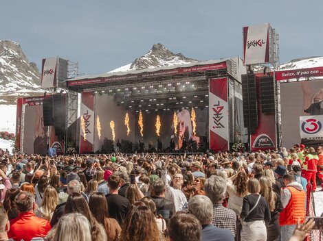 Ein großes Musikfestival in den Bergen mit einer beeindruckenden Bühne. Viele Menschen genießen die Aufführung unter klarem Himmel.