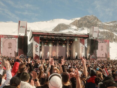 Ein großes Musikfestival in den Bergen mit vielen Menschen, die zur Show tanzen. Im Hintergrund sind schneebedeckte Berge und eine Bühne mit Lichtern zu sehen.
