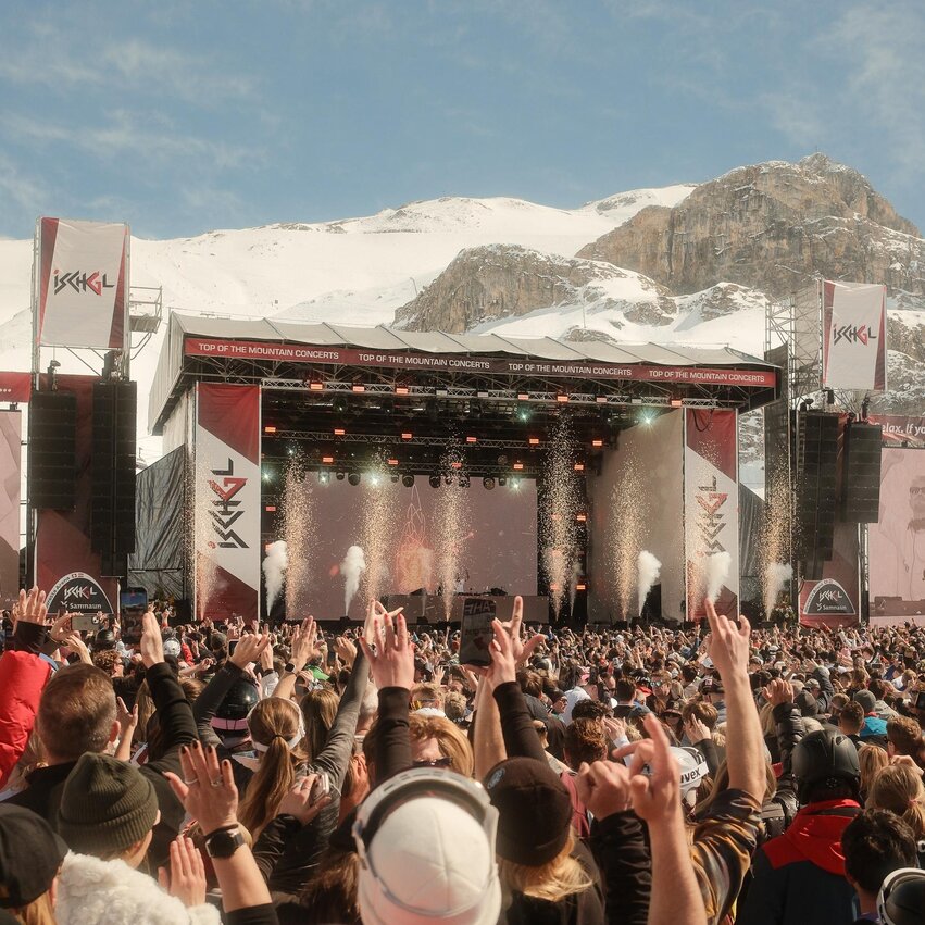 Ein großes Musikfestival in den Bergen mit vielen Menschen, die zur Show tanzen. Im Hintergrund sind schneebedeckte Berge und eine Bühne mit Lichtern zu sehen.
