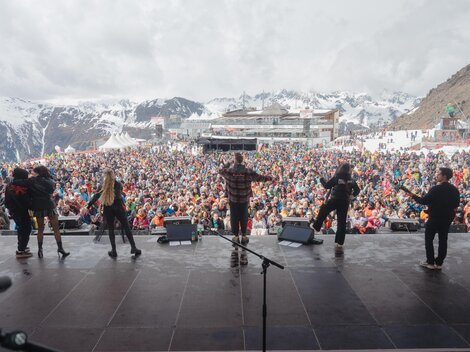 Eine große Menschenmenge genießt ein Konzert in den Bergen. Im Hintergrund sind schneebedeckte Gipfel und ein Veranstaltungsgebäude zu sehen.