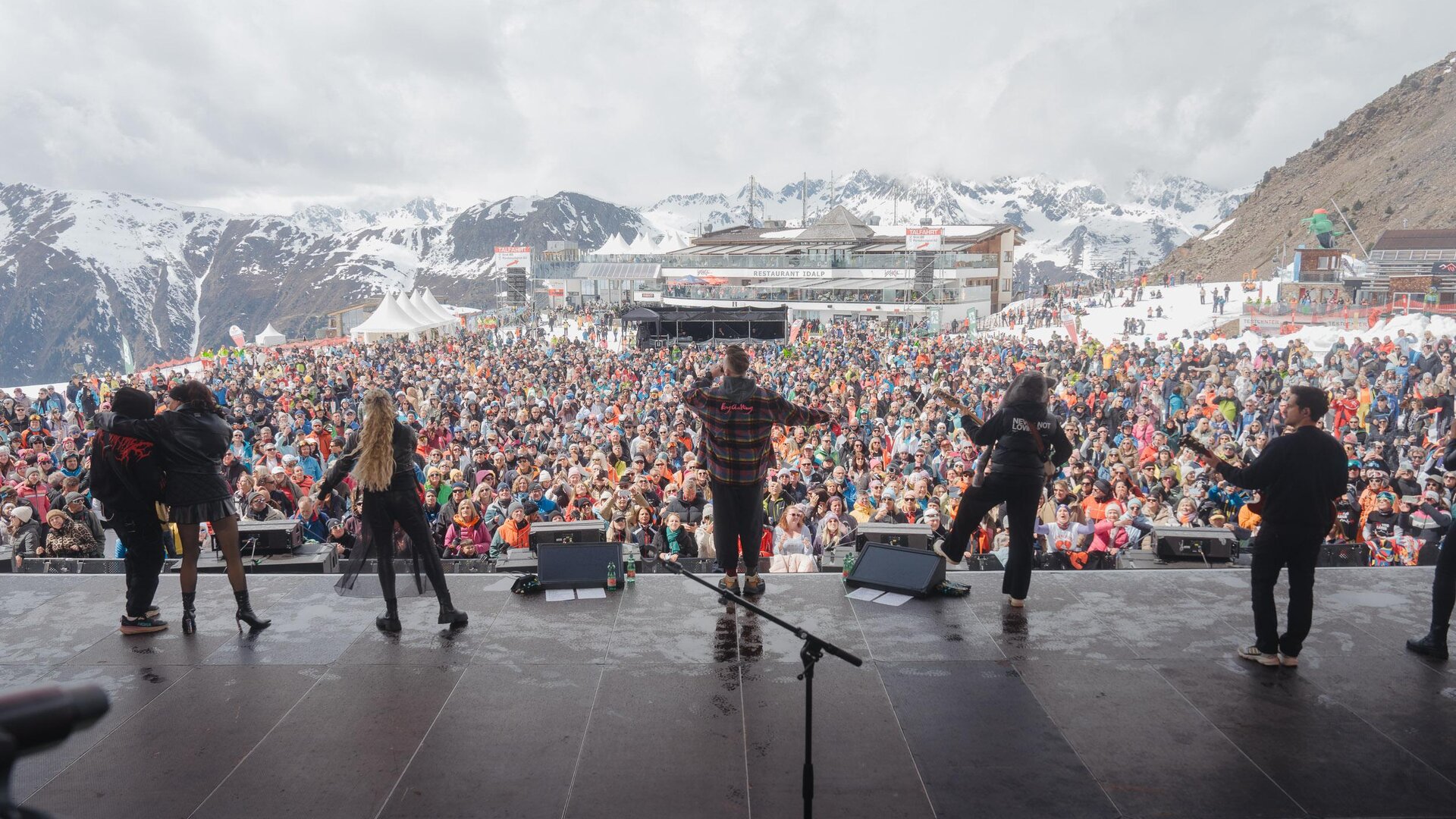Eine große Menschenmenge genießt ein Konzert in den Bergen. Im Hintergrund sind schneebedeckte Gipfel und ein Veranstaltungsgebäude zu sehen.
