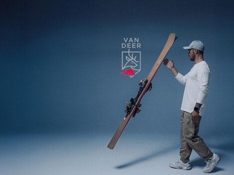 A man is holding skis in a calm, blue environment. In the background, there is a sign for a pop-up testing center in Ischgl from December 5 to 8, 2025.