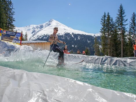 A skier is going over a water jump in an alpine setting. In the background, snow-covered mountains and pine trees can be seen.