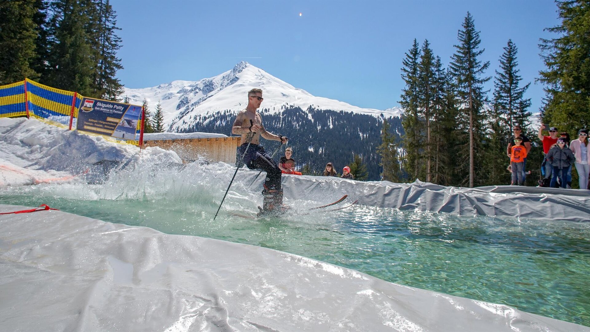 A skier is going over a water jump in an alpine setting. In the background, snow-covered mountains and pine trees can be seen.