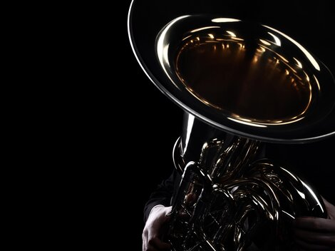 A musician holds a shiny tuba in the dark. Only the lower part of the instrument and the hands are visible.