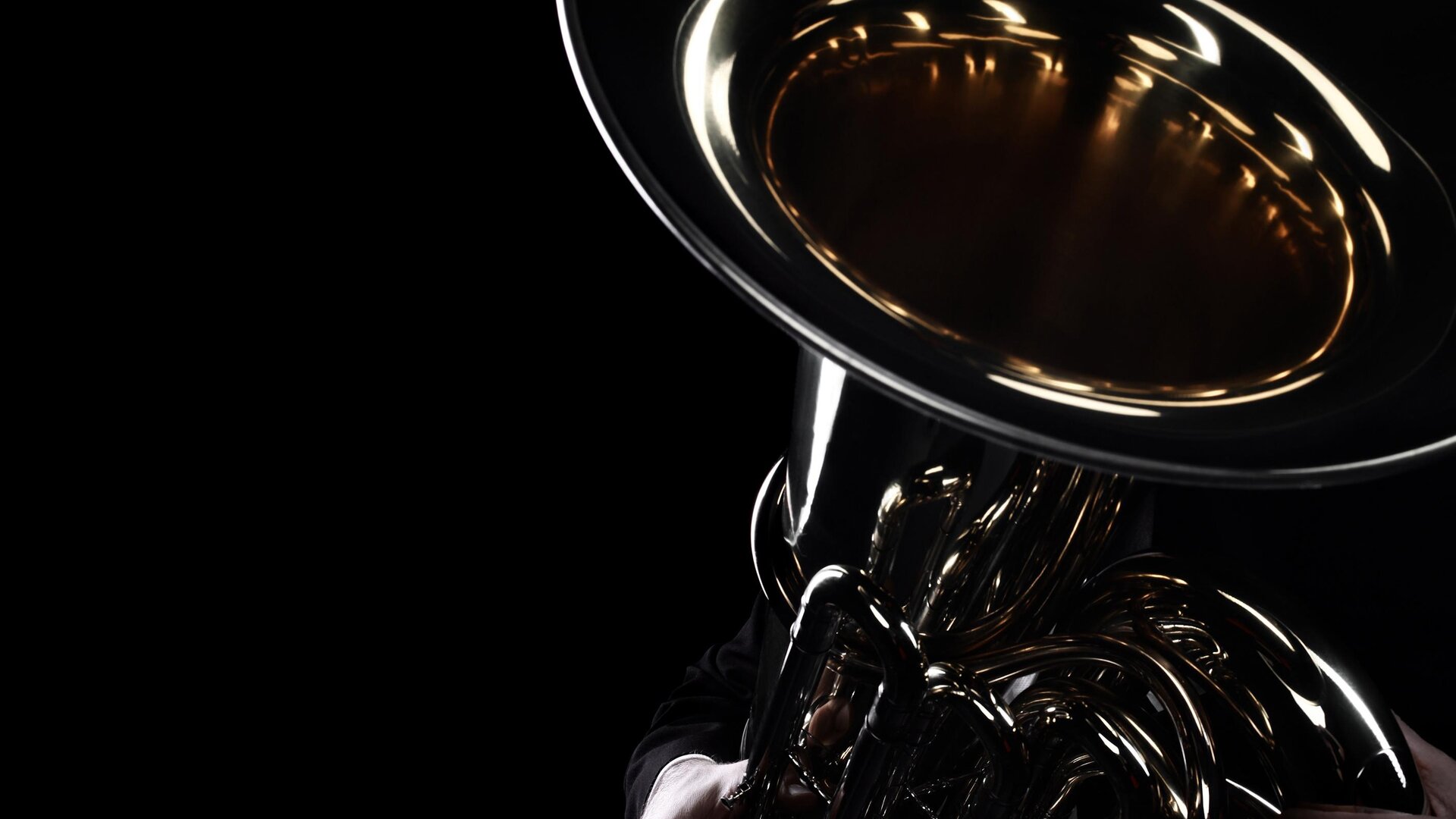 Ein Musiker hält eine glänzende Tuba im Dunkeln. Nur der untere Teil des Instruments und die Hände sind sichtbar.
