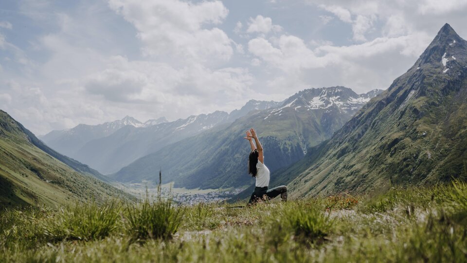 Eine Person übt Yoga in einer malerischen Berglandschaft. Die Umgebung ist grün und die Berge sind teilweise schneebedeckt, unter einem klaren Himmel.