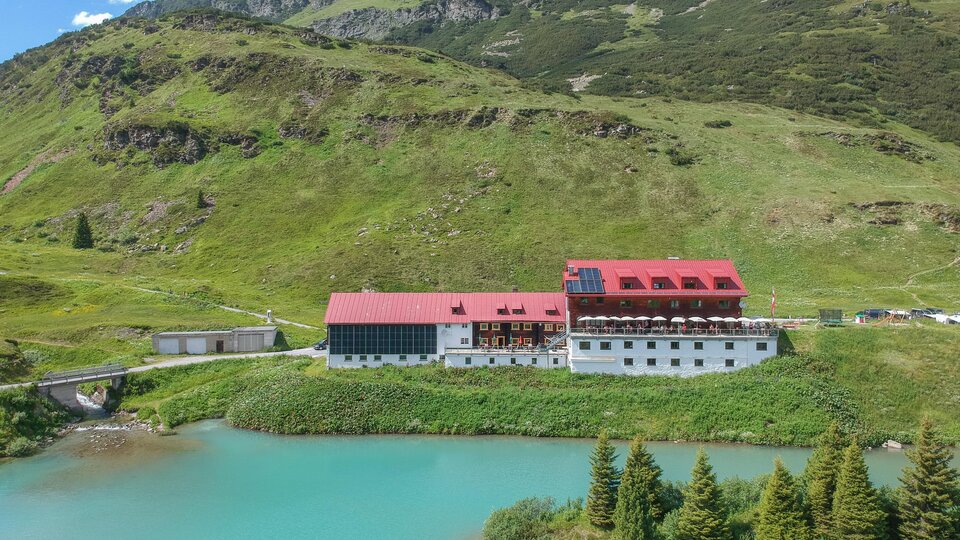 A picturesque hotel by the shore of a clear, blue lake, surrounded by green hills. In the background, mountains and a blue sky can be seen.
