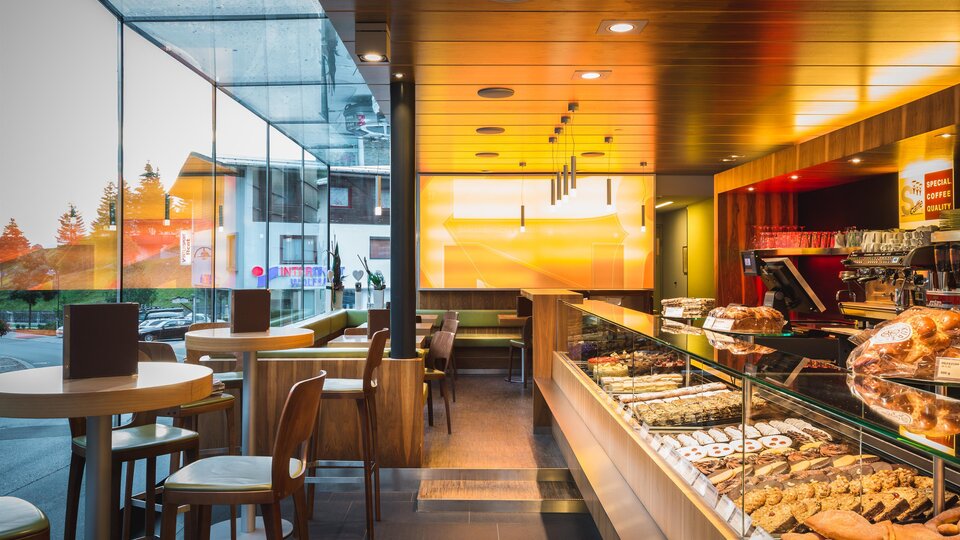 A modern café with a large window front and inviting atmosphere. The counter is decorated with various pastries and drinks.