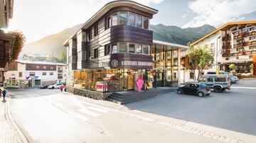 A modern building with large windows and appealing architecture. In the background, mountains and a clear sky are visible.