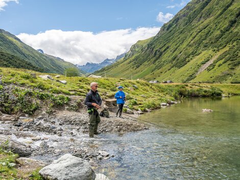 Zwei Angler stehen am Ufer eines klaren Gewässers und fischen. Die Umgebung ist grün und einladend.