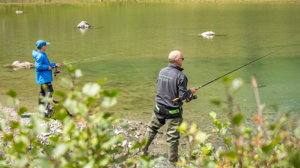 Zwei Angler stehen am Ufer eines klaren Gewässers. Im Hintergrund sind grüne Wiesen und Steine zu sehen.
