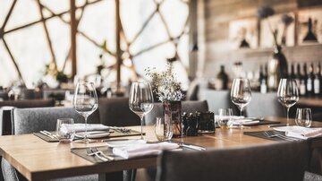 Ein elegantes Restaurant mit stilvollem Tischarrangement. Gläser und Besteck sind vorbereitet und eine Blumendekoration verleiht dem Ambiente eine einladende Note.