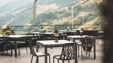 Eine Terrasse mit mehreren Tischen und Stühlen, umgeben von Berglandschaft. Die Atmosphäre ist einladend und ruhig.